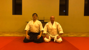 Two men, kneeling on a mat. They appear to be practicing a form of martial arts.