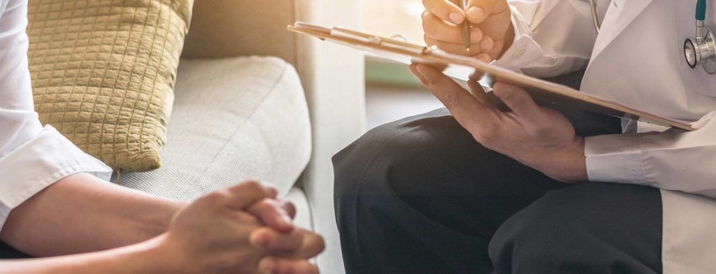 A doctor, across from an individual, writing on a clipboard.