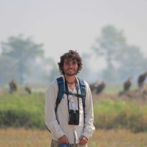 Dan, shown on his travels, in a white shirt. He has binoculars and is in an agricultural field.