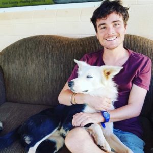 Dan, with a dog. He is sitting on a couch and wearing a maroon t-shirt.