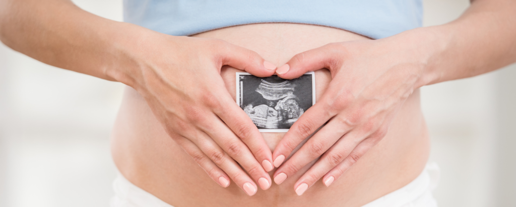 Hands in the shape of a heart, resting on a belly. Within the heart is a printed ultrasound image.
