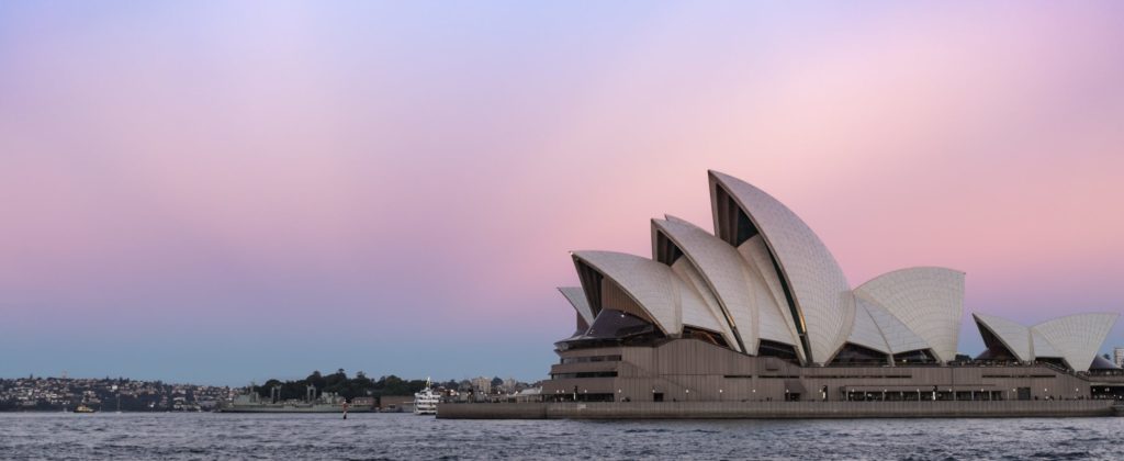 The Sydney Opera House, pictured from across the harbour. The sky is tinted pink and purple.