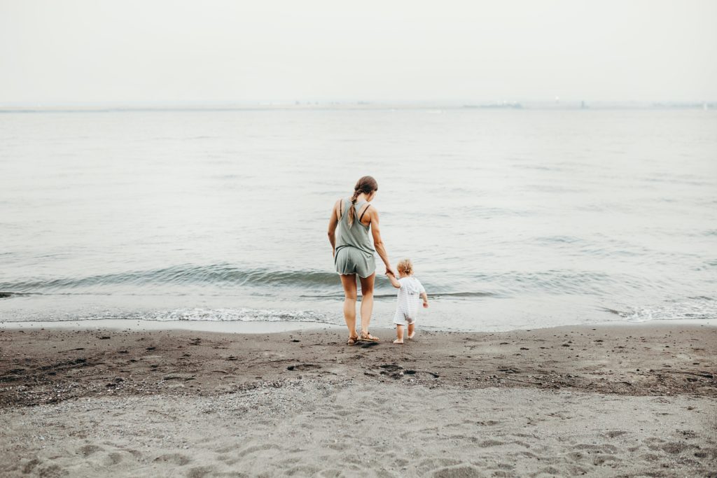 A woman and her child, holding hands and walking towards the ocean. They are shown from behind.