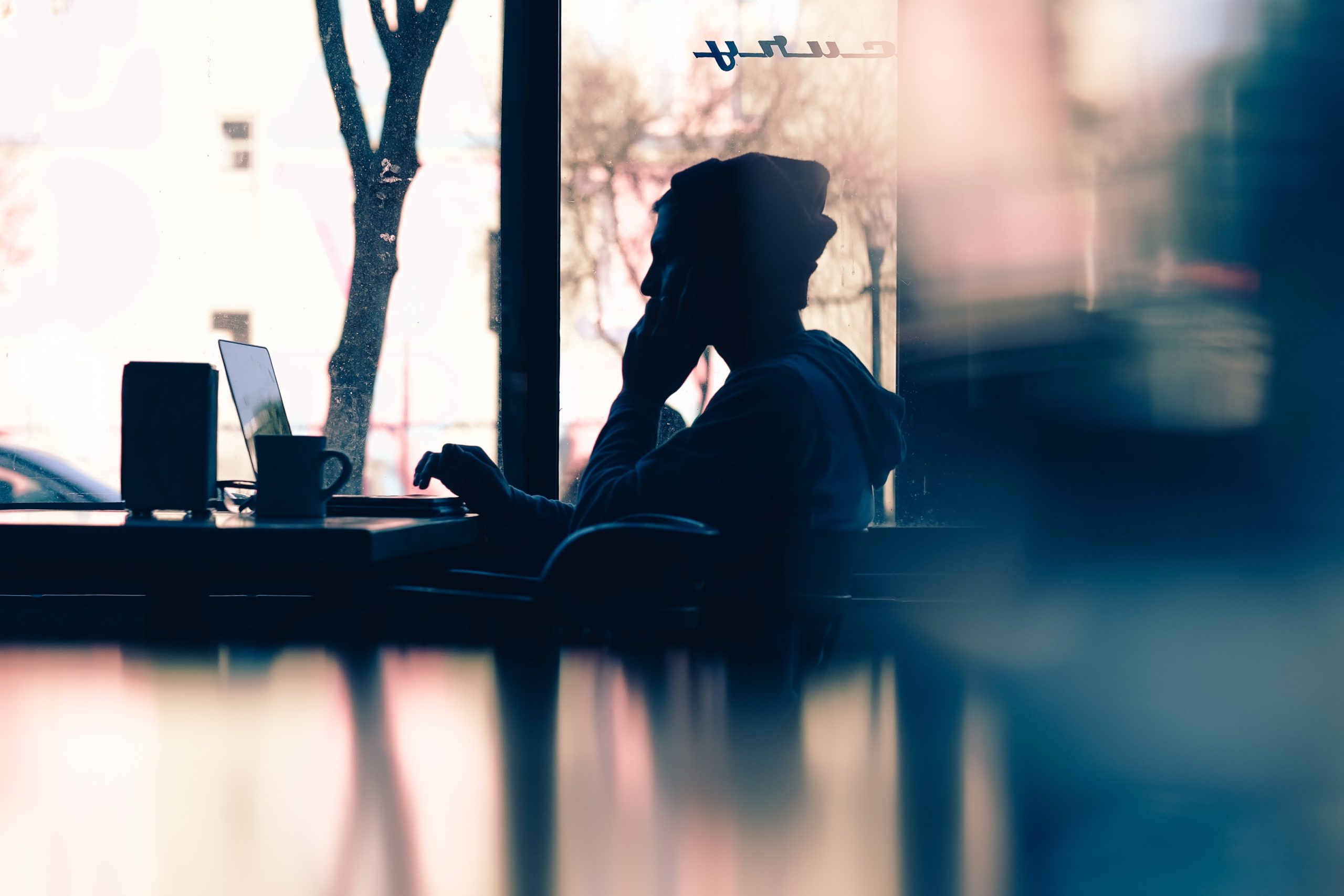 An individual, shown side on, talking on the phone whilst sitting at a table with their laptop open in front of them.