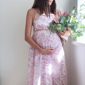 Alex wearing a white and pink floral dress, and holding her pregnant belly in one hand. She is holding a bouquet in the other hand.