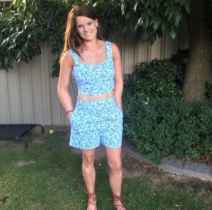 Tash, standing in a backyard and smiling at the camera. She is wearing a blue outfit.