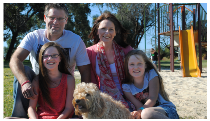 A family of four and their dog at the park. They are crouching down and smiling at the camera.