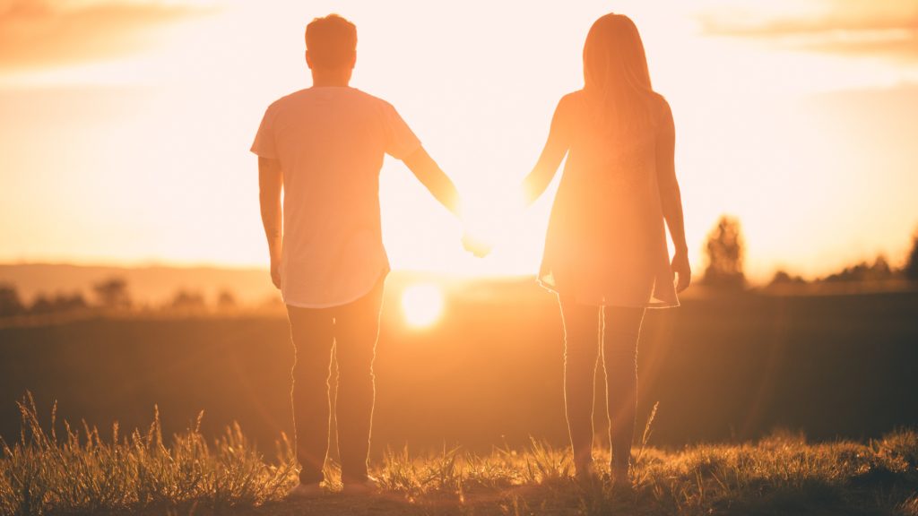 Two people holding hands with their backs to the camera. They are outside on a hill, and the sun is setting in front of them, casting an orange glow over the image.