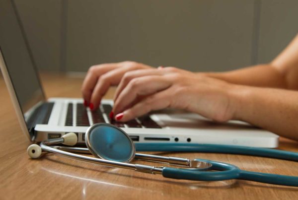 Hands typing on a laptop on a table. There is a stethoscope next to the laptop.