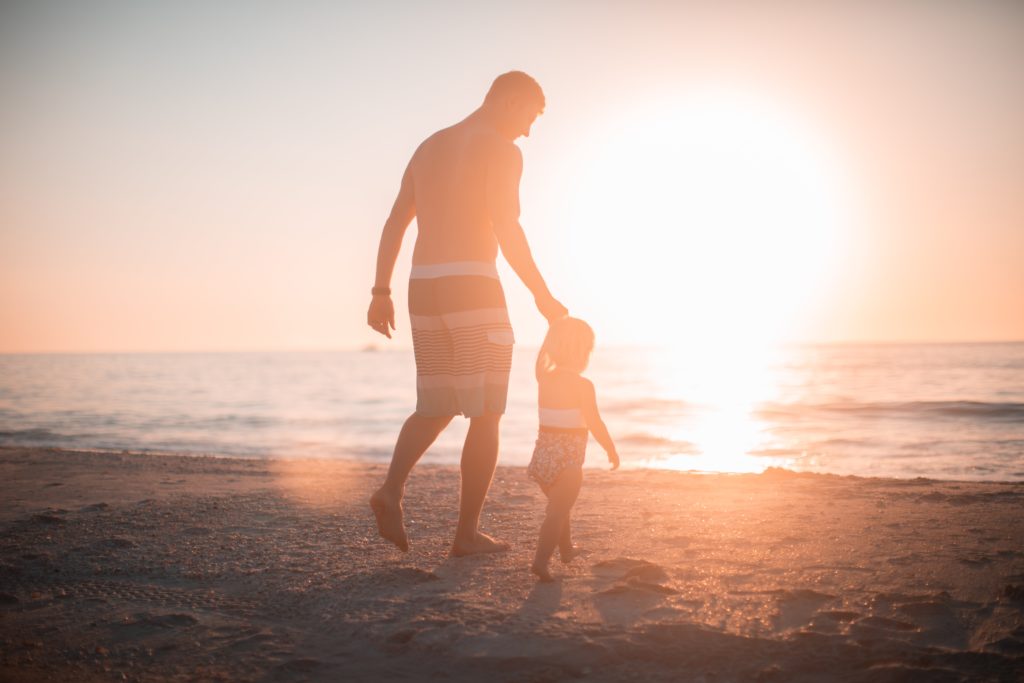 A man and a young child walking towards the ocean at the beach. They are holding hands, and the sun is setting in front of them.
