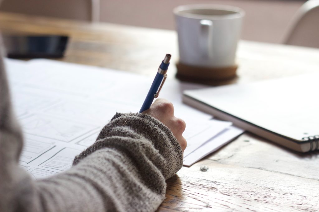 An individual writing with pen and paper. Their hand is in the foreground and a notebook and mug are in the background.
