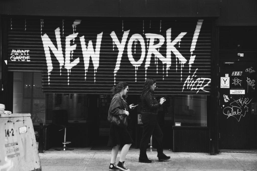 A greyscale photograph of two women walking in front of a a piece of street art showing the words New York.