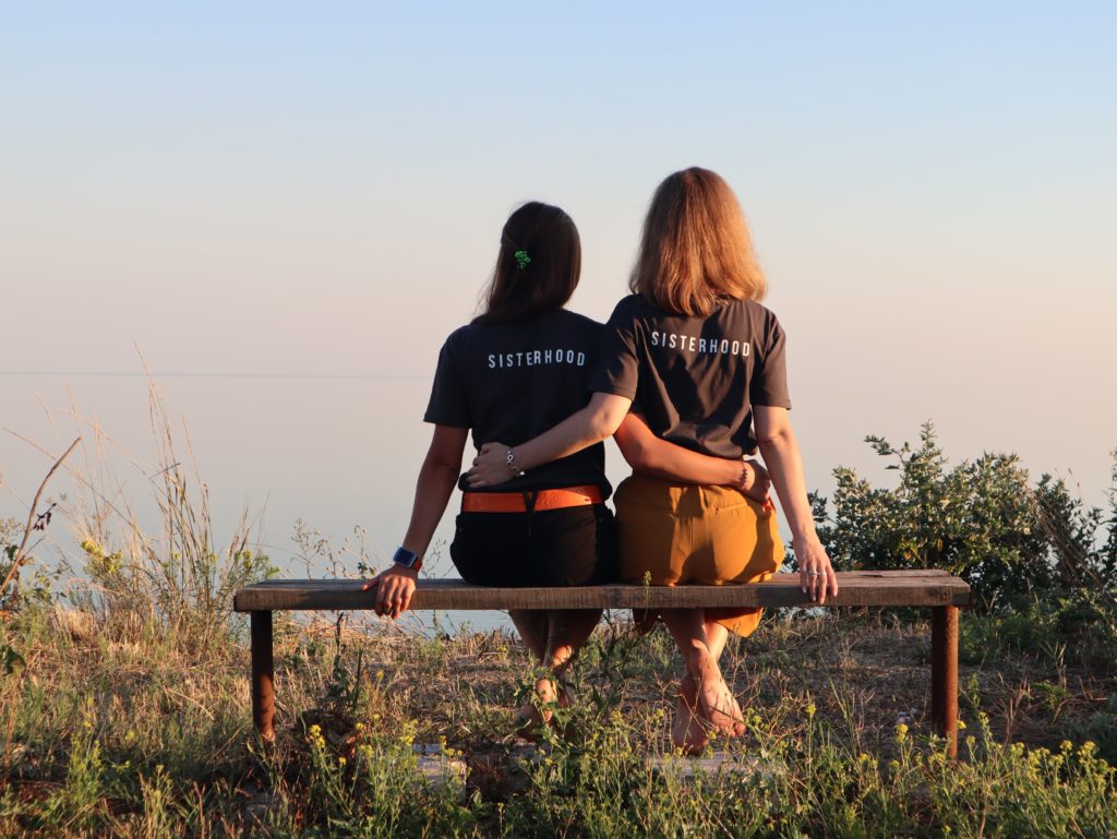 CF specific resources for young people | Two women sitting on a bench, backs to the camera. They are looking at the natural landscape ahead of them, and have their arms around each other. The backs of both of their t-shirts say Sisterhood.