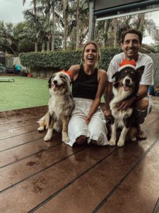 Jess, sitting on a deck with two dogs and a man, smiling at the camera.
