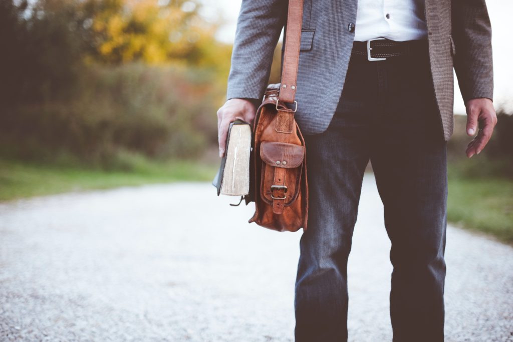 A photograph of a man from the chest down. He is wearing a suit and holding a book. He also has a brown leather briefcase over his shoulder.