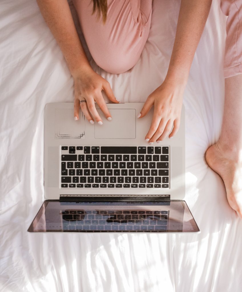 A birds eye view of a women sitting on a bed, using a laptop. She is wearing pink pants.