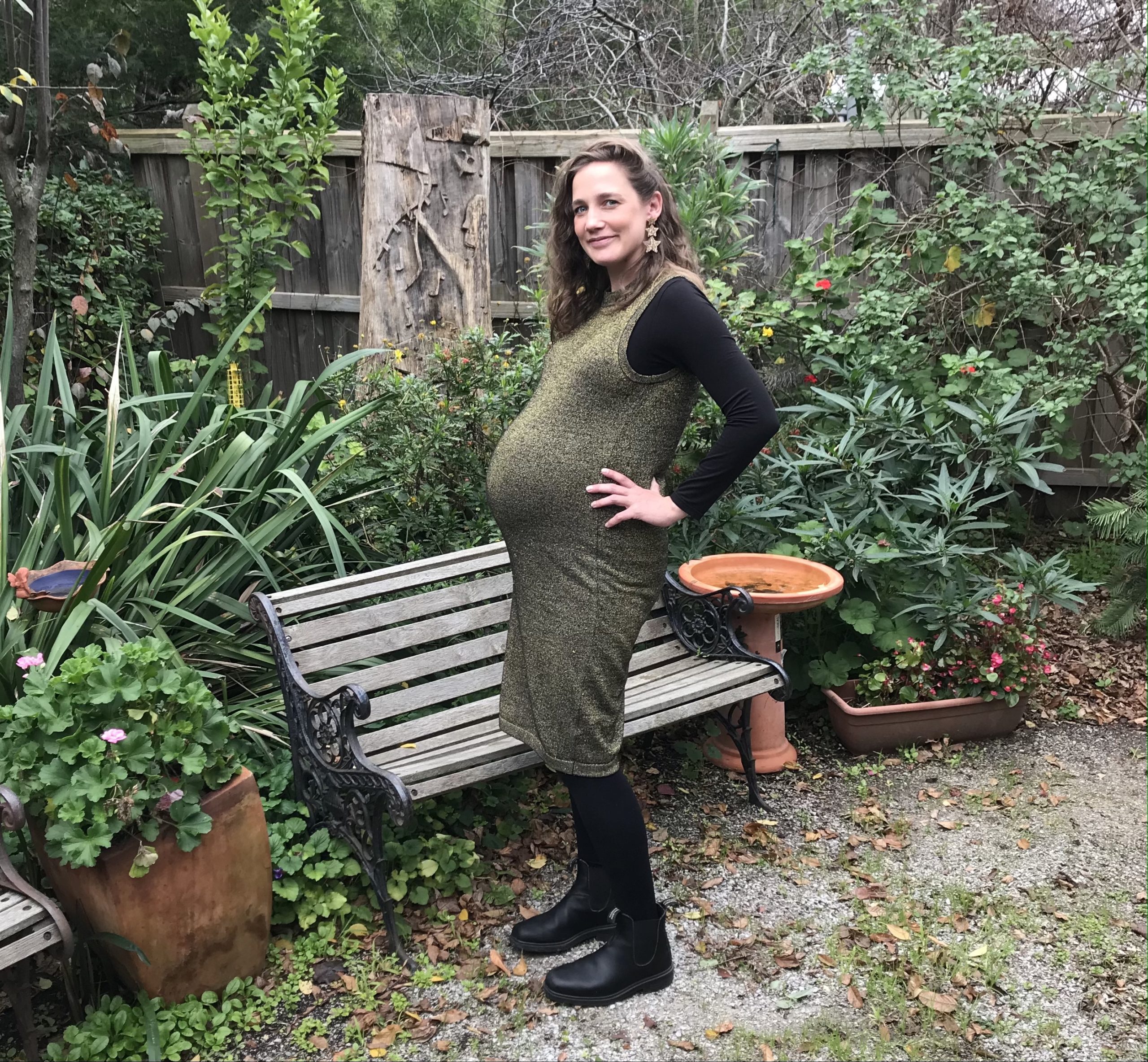 Emily standing in front of a bench in a lush garden. She is pregnant and has her hands on her hips.