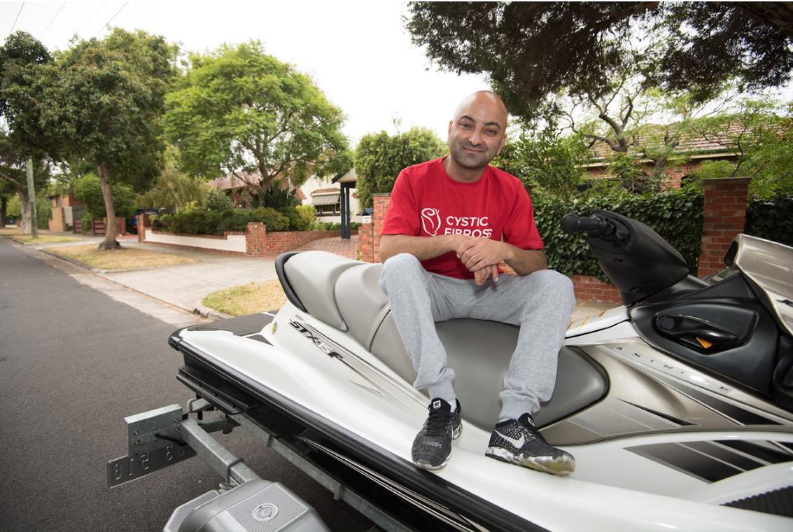 Tony, sitting on a jet ski on a trailer. He is wearing a red t-shirt with Cystic Fibrosis written on it in white.