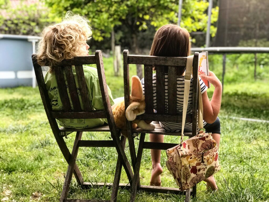 Two children, sitting outside on wooden chairs, backs to the camera. One child is reading a book, and there is a soft toy in between the chairs.