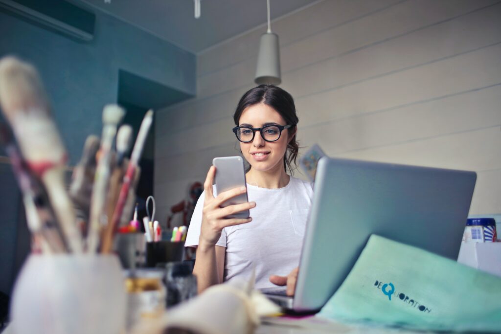 A woman with glasses sitting in front of a laptop, looking at her phone. There are blurred out paintbrushes in the foreground.