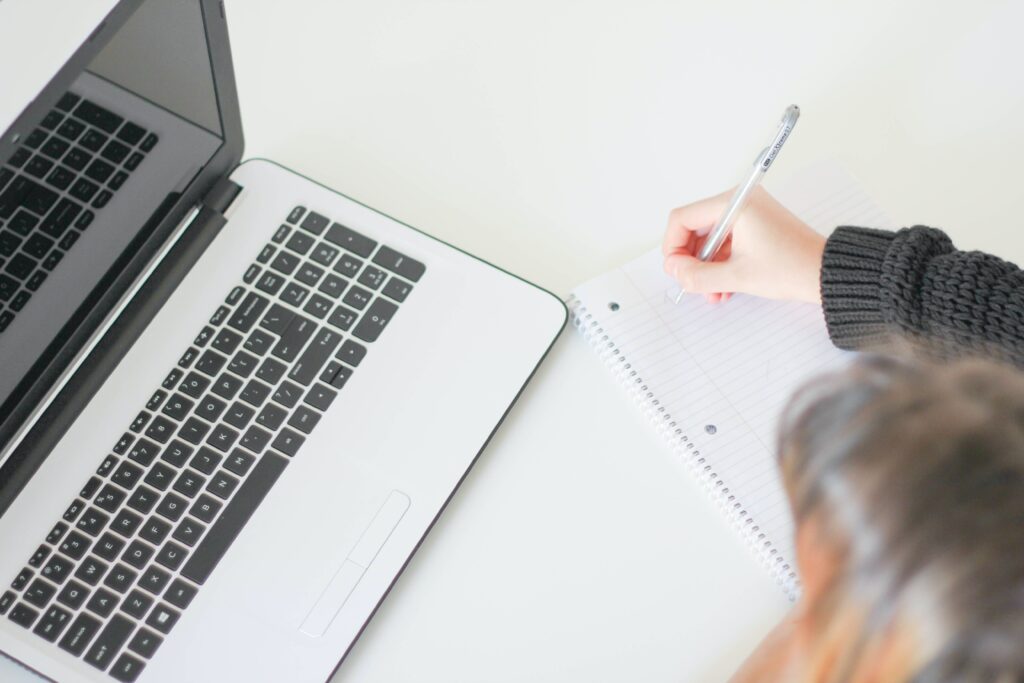 A birds-eye view of an individual writing down notes into a notebook, with a laptop open in front of them.