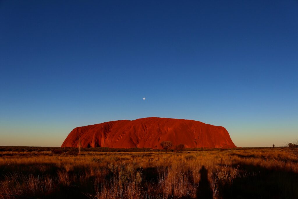 Uluru, pictured under a clear yellow and blue sky, at sunset or sunrise.