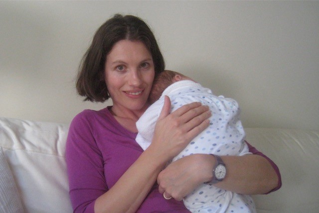 Serena, holding a newborn child on a couch. Serena is smiling at the camera.