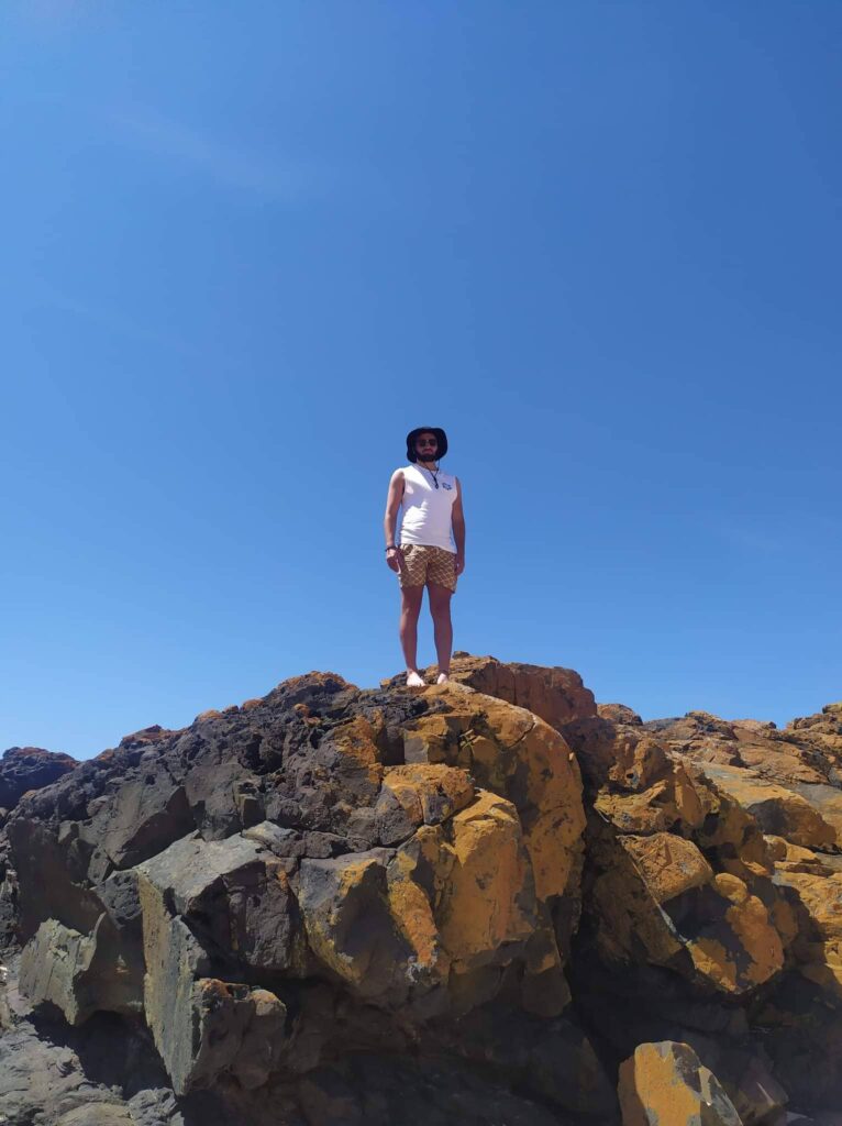 Sam Lefoe, standing on a large rock on a sunny day. He is wearing a hat and sunglasses.