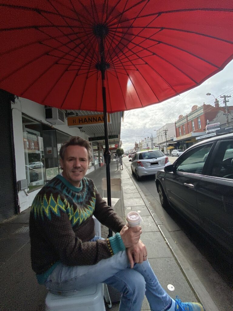 James sitting on a bench, under a red umbrella. He is holding a takeaway coffee.