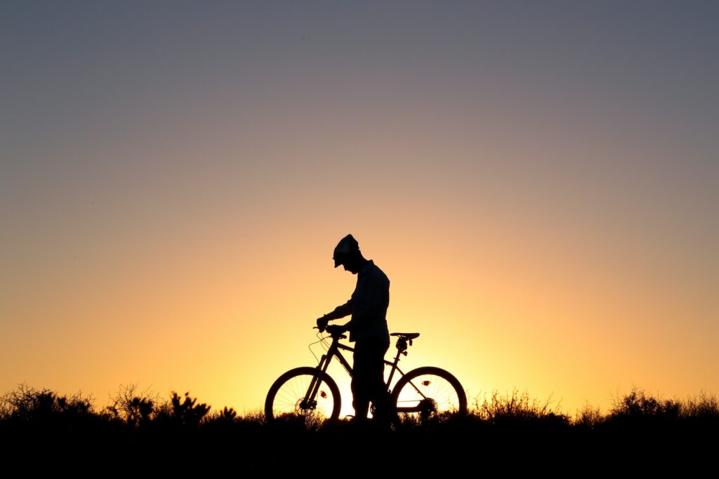 Image is of a person standing next to a bike in silhouette against a sunrise. The person and bike are black against the brightness of the sunrise.