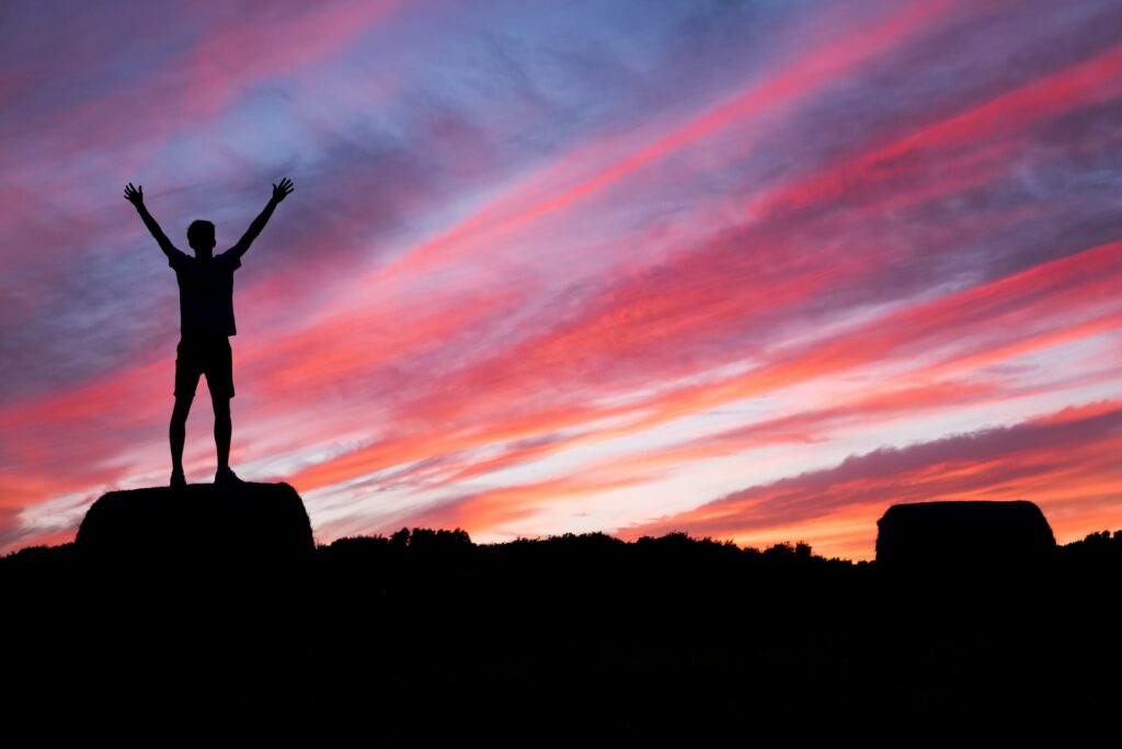 A silhouette of an individual standing with their arms in the air, against a sky at sunset, coloured purple, pink and orange.