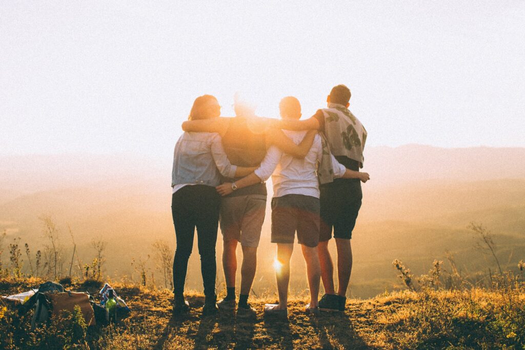 Social Wellbeing | A group of people standing on a hill, with their arms around each other and backs to the camera. The rising/setting sun casts a yellow glow over the image.