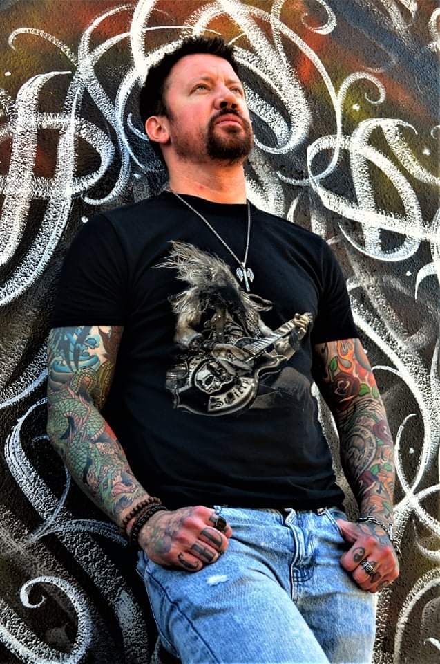A man leaning on a wall and looking upwards. He has a necklace and tattoo sleeves on his arms.