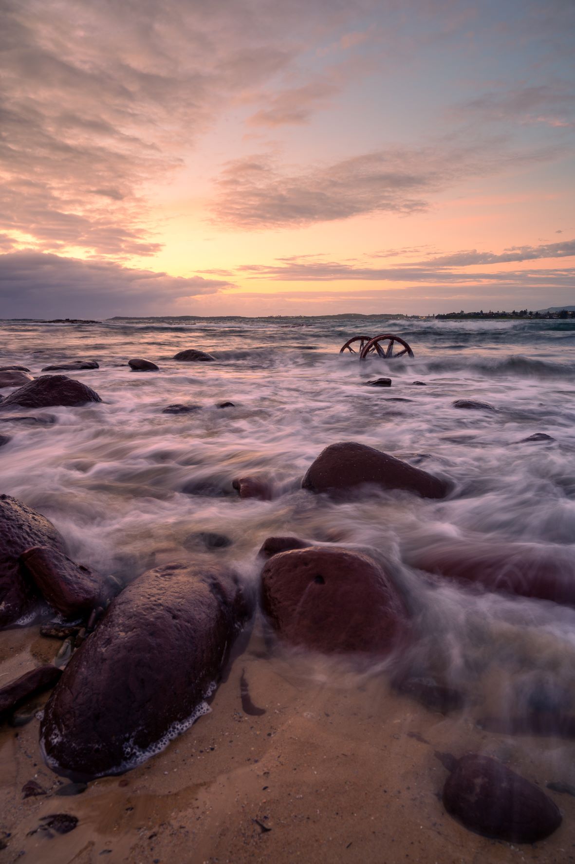 Image by Rory O'Keefe. Photo is of the beach at sunrise. In the foreground, water spills over rocks and sand. In the background, the sun rises against a pale blue and yellow sky. A smattering of clouds litter the sky.