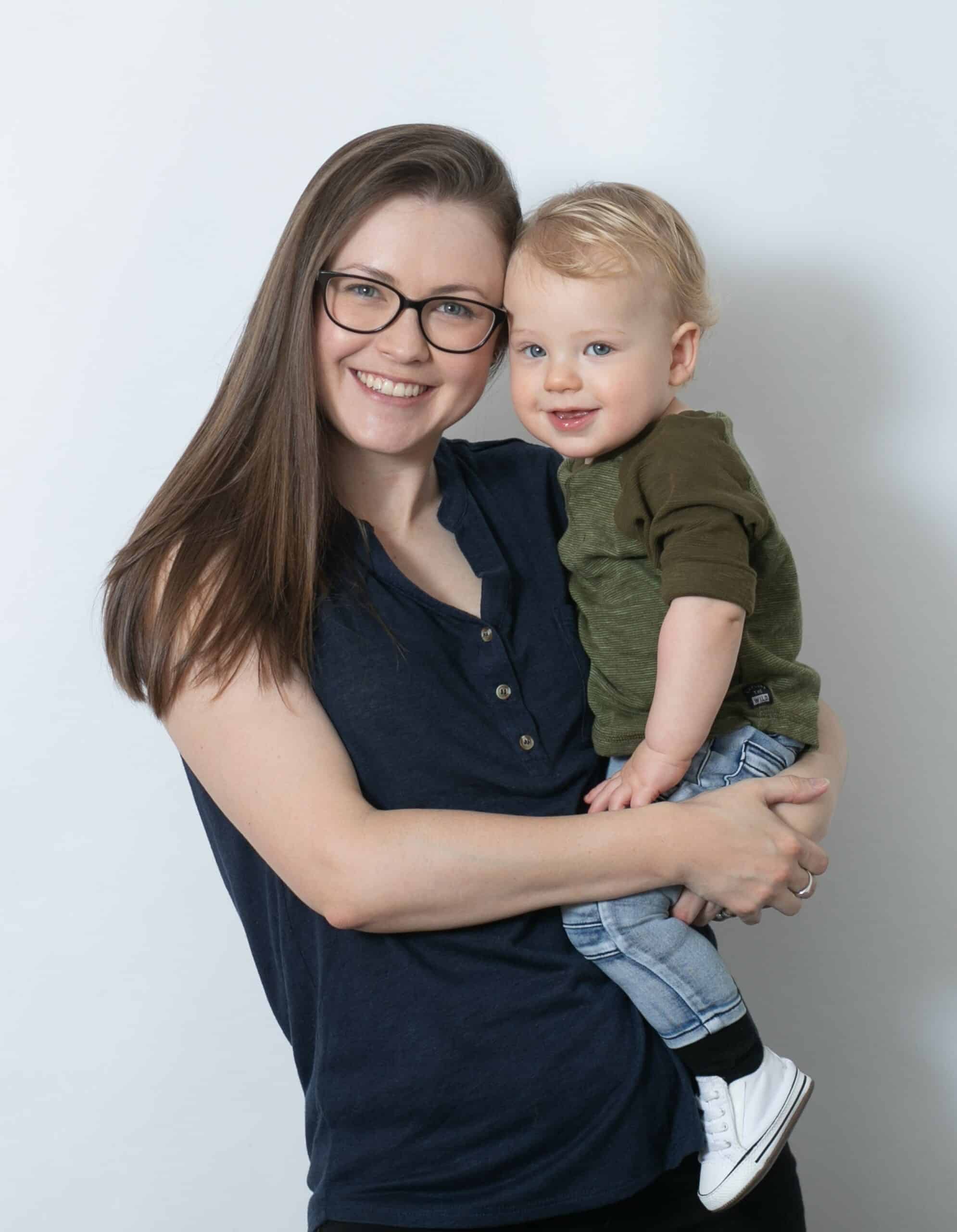 Image is of a woman wearing glasses and with long brown hair holding a baby boy. They are both looking towrads the camera and smiling.