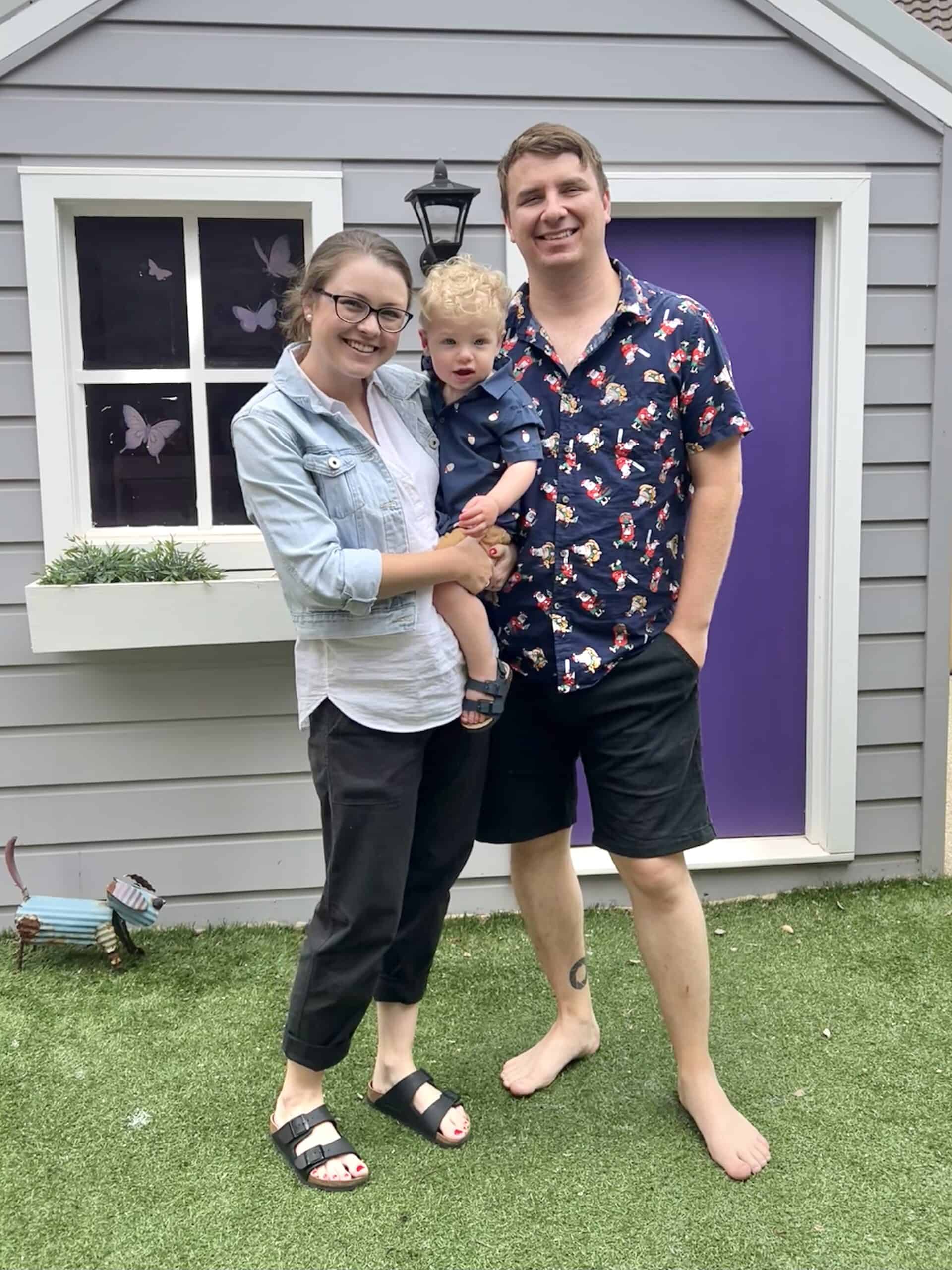 Image is of two adults (one woman and one man) and a baby boy standing in front of a grey and white cubby house with a purple door. The woman holds the baby boy. All three are looking towards the camera and smiling.