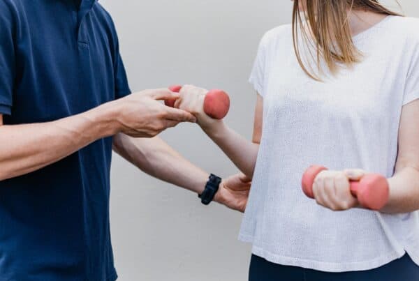 Two people stand next to each other. One, a white woman, holds two small pink weights in her hands. The other person, a white man, rests a hand on her elbow nd the other on her fist around the weight. He is providing guidance and support.
